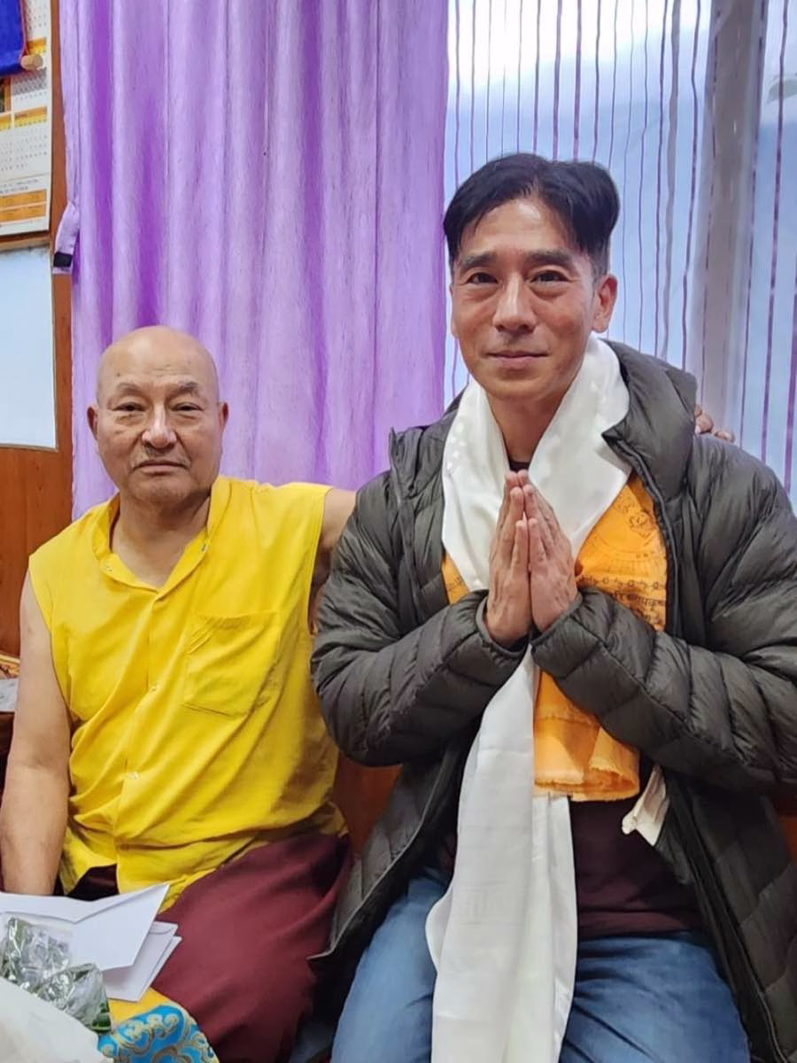 Alex Yamane with palms joined, wearing a white khata scarf, beside a Buddhist lama in yellow robes